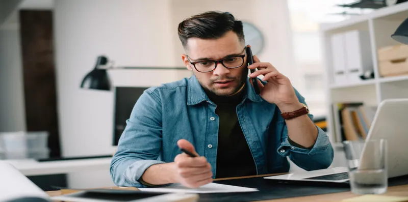 man sitting at desk with laptop and papers looking concerned talking on the phone