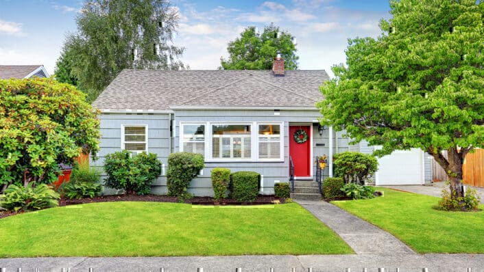 Pequeña casa azul con puerta roja y jardín con plantas de hoja perenne.