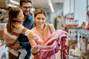 Little girl and her parents choosing backpack for school while shopping in store together.